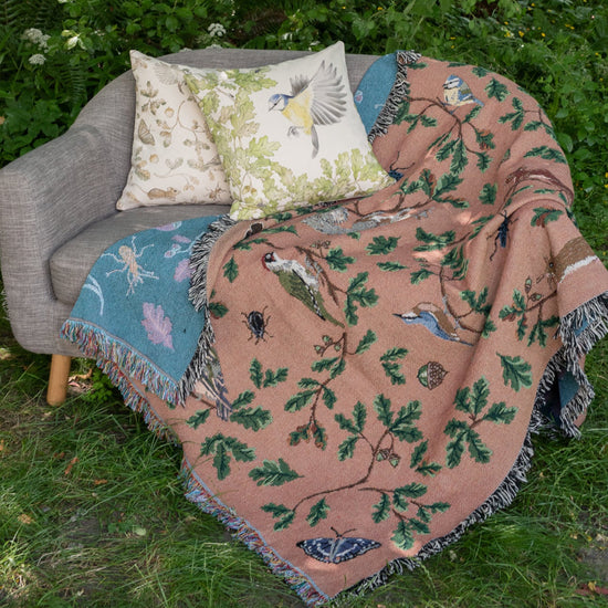 russet coloured woven blanket with teal reverse featuring birds, squirrels and oak, draped over sofa in garden