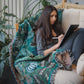 woven wrap blanket with fringe featuring lots of different houseplants draped over chair surrounded by houseplants with woman sitting and reading