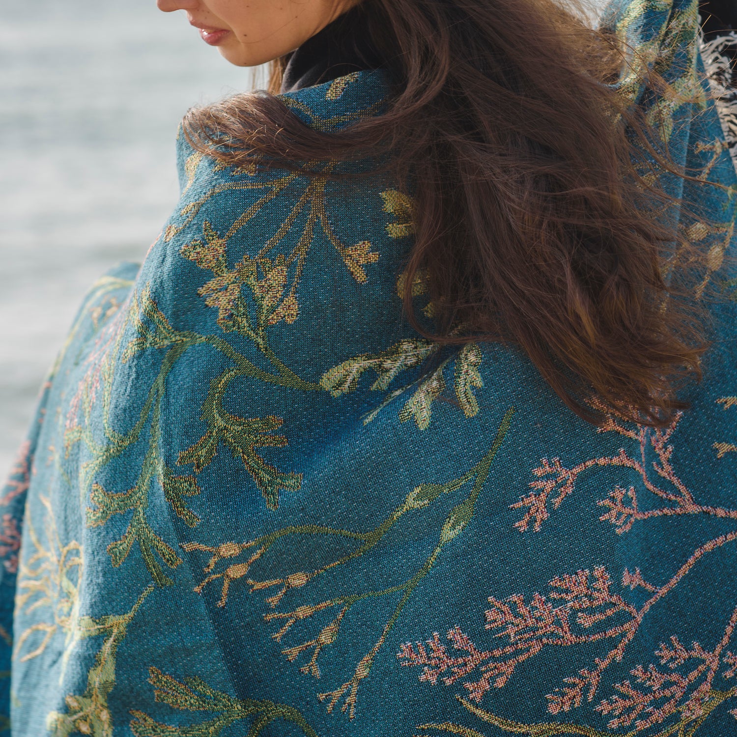 delicate seaweed design on woven wrap blanket with fringe wrapped around womans shoulders with the sea in the background