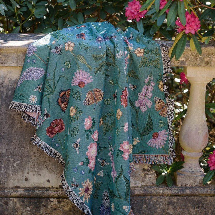 Pollination Bloom woven blanket draped over stone wall in afternoon sunlight showing butterflies and flowers detail