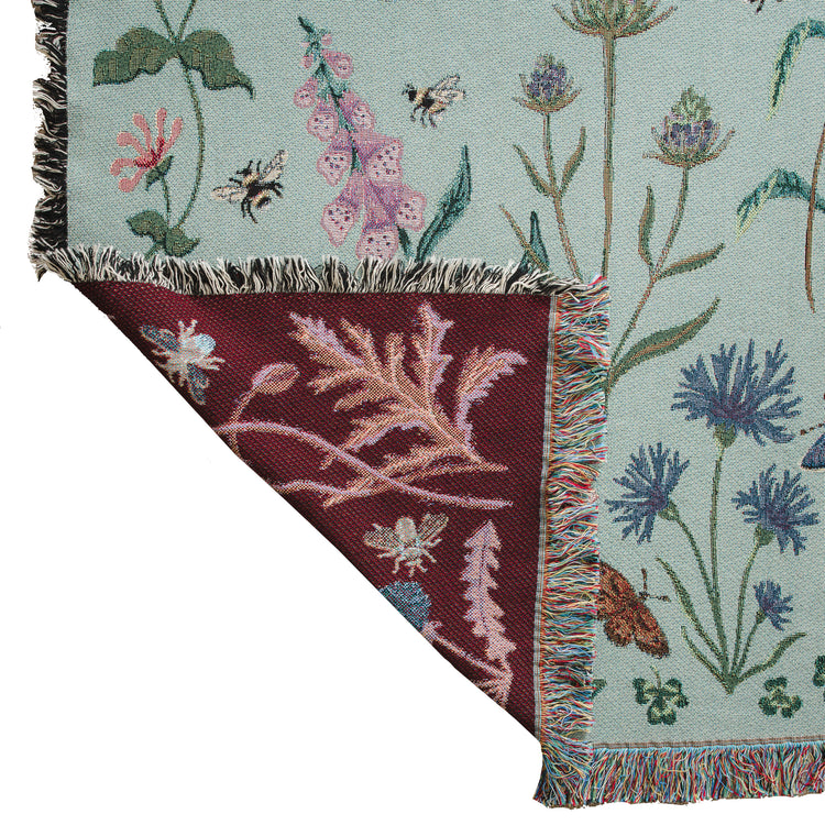 pale sage green woven blanket with ruby red reverse with details of butterflies and wild flowers, showing fringe and woven details
