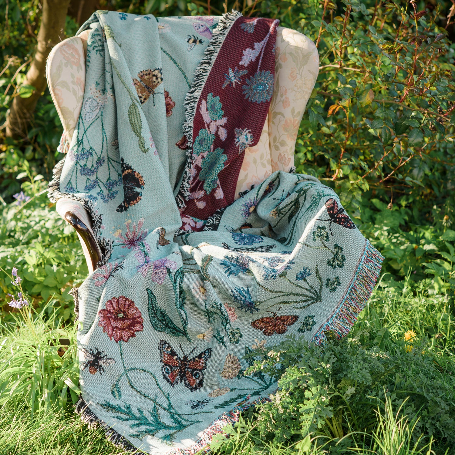 pale sage green woven blanket with ruby red reverse with details of butterflies and wild flowers, draped over armchair in summer garden