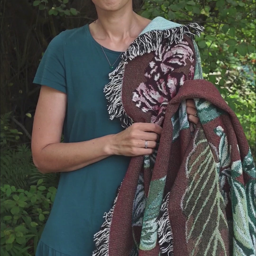 Video showing woven throw blanket featuring houseplants warm burgundy tones on the front, contrasted with light aquamarine on the back.