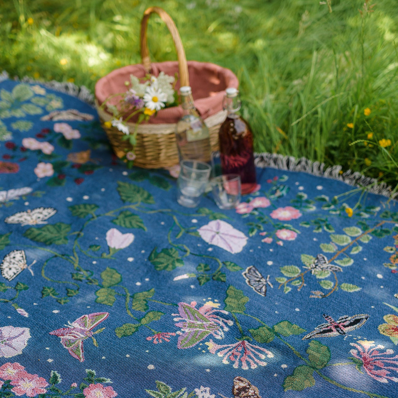 deep blue woven blanket with intricate moth and flower design being used as a magical picnic blanket with drinks and picnic hamper