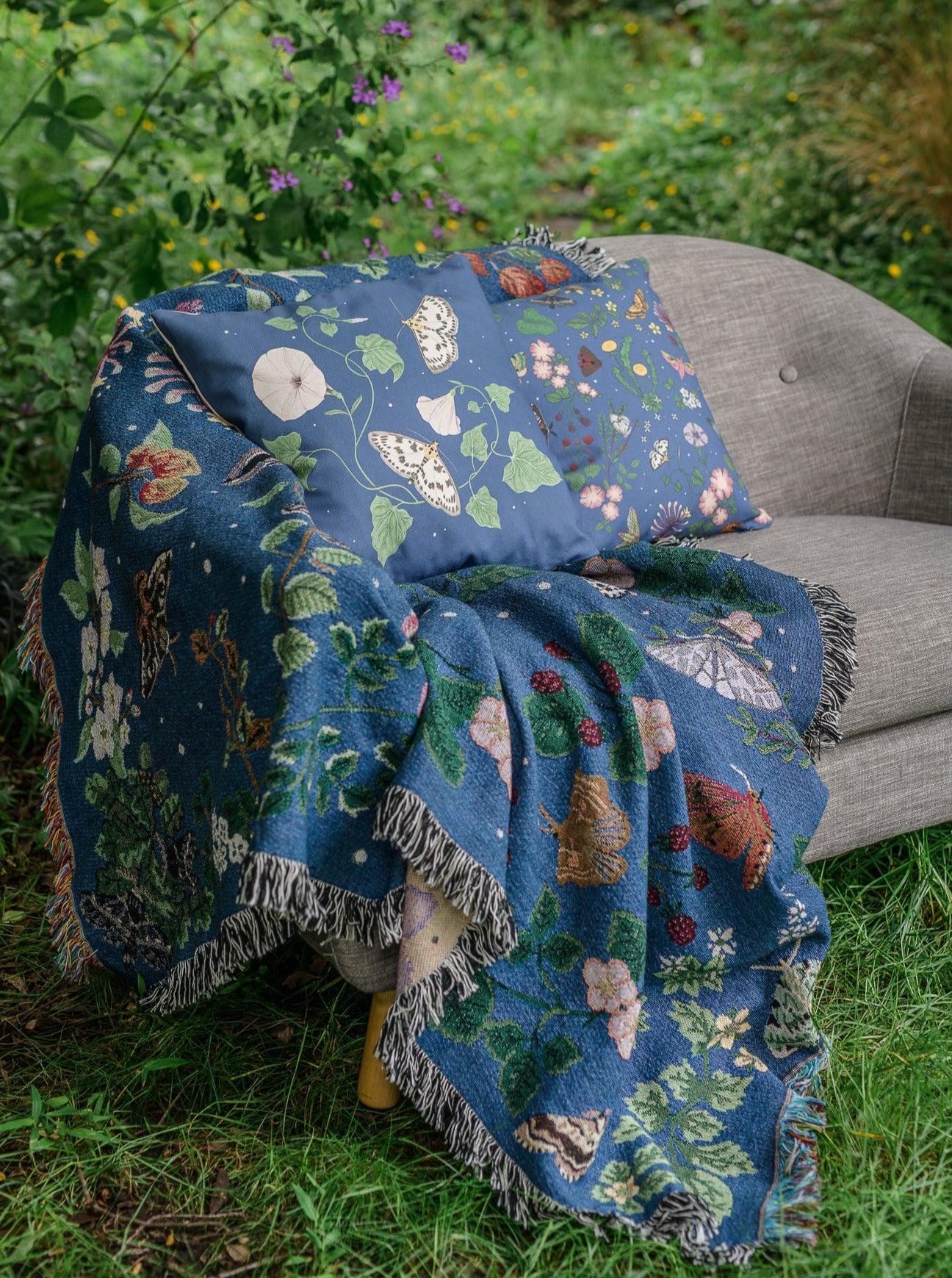 deep blue woven blanket with intricate moth and flower design being thrown across a grey sofa in a wild garden