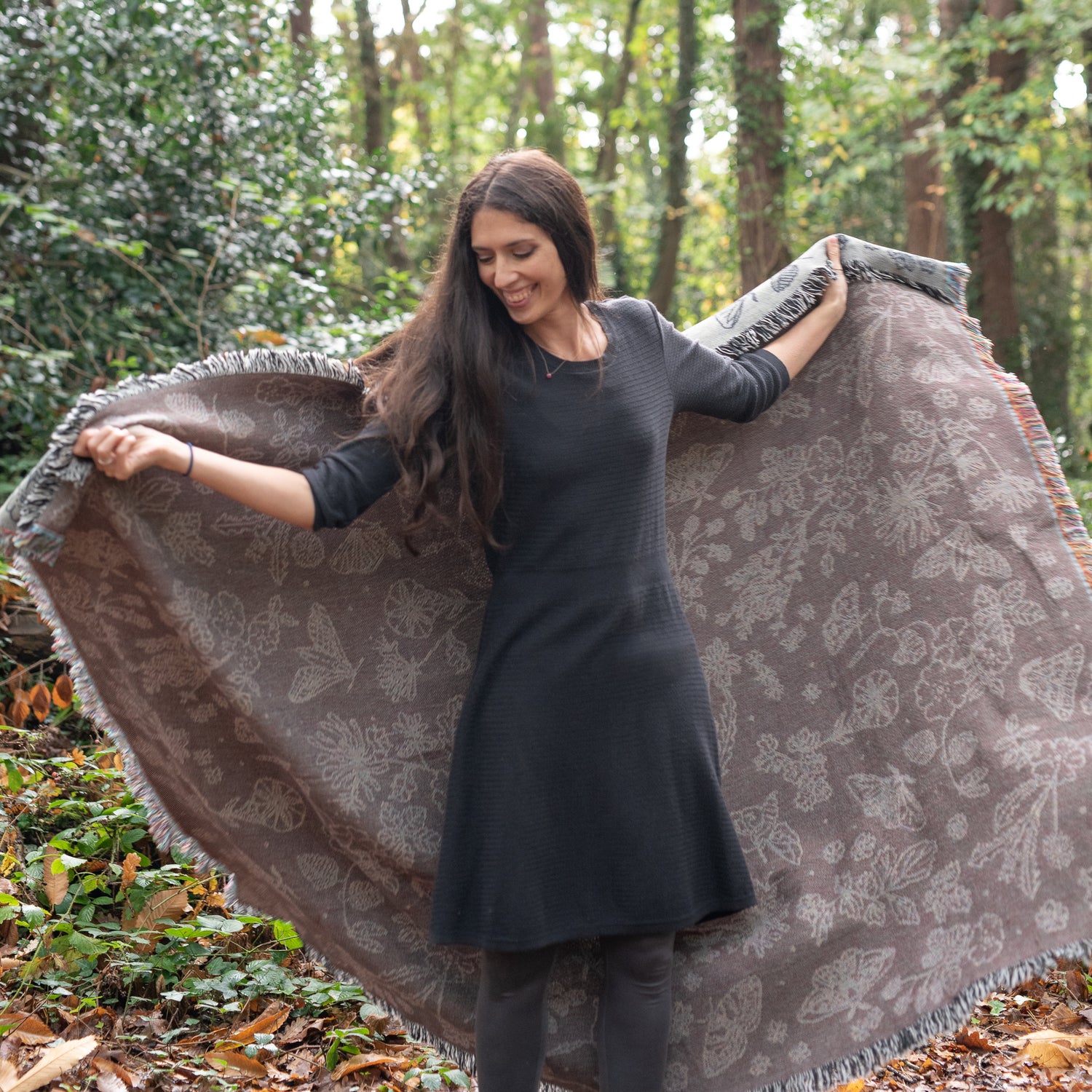 Pretty grey blanket with black pattern of moths, berries, leaves and flowers being held up by happy woman in the woods