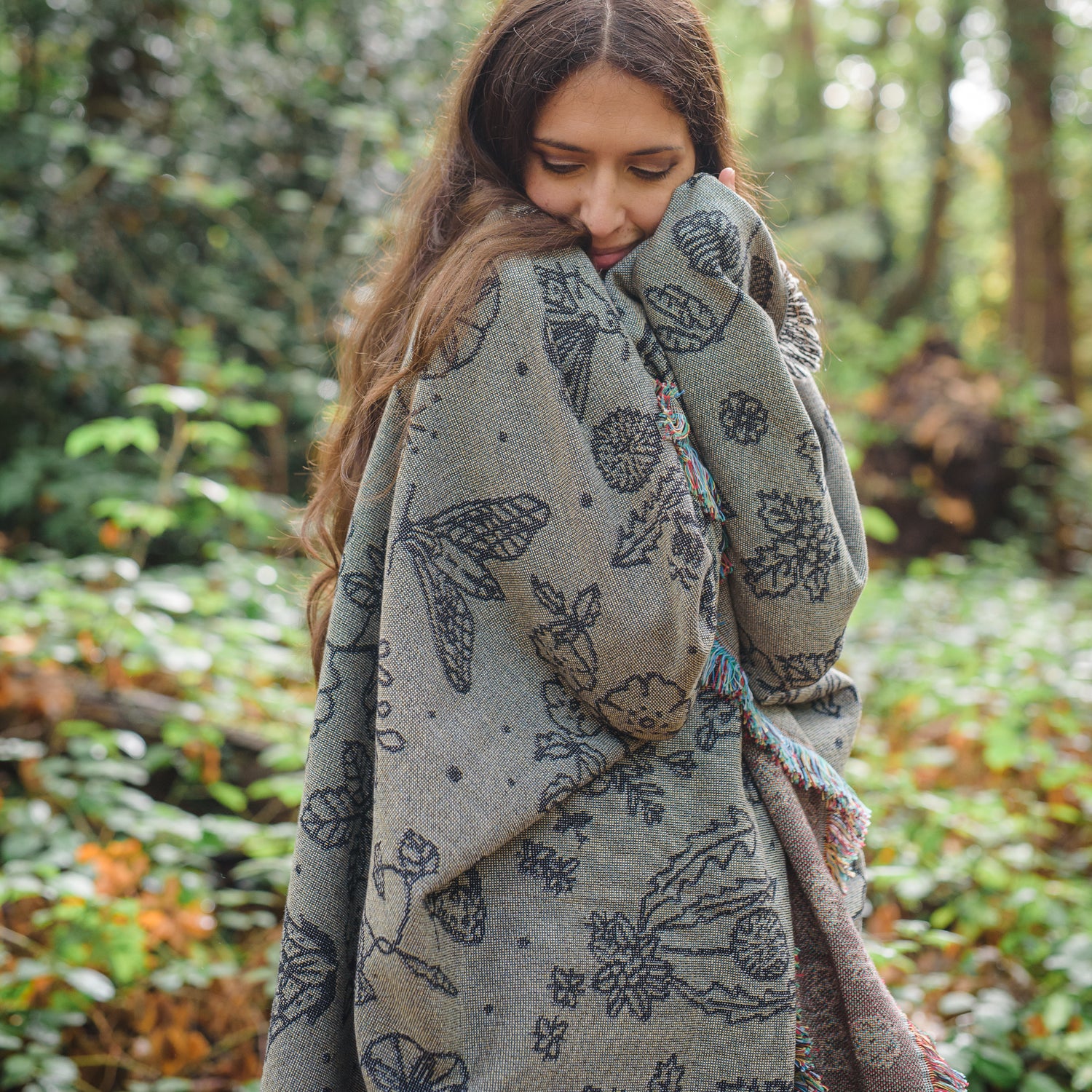 Pretty grey blanket with black pattern of moths, berries, leaves and flowers wraped around a woman in the woods
