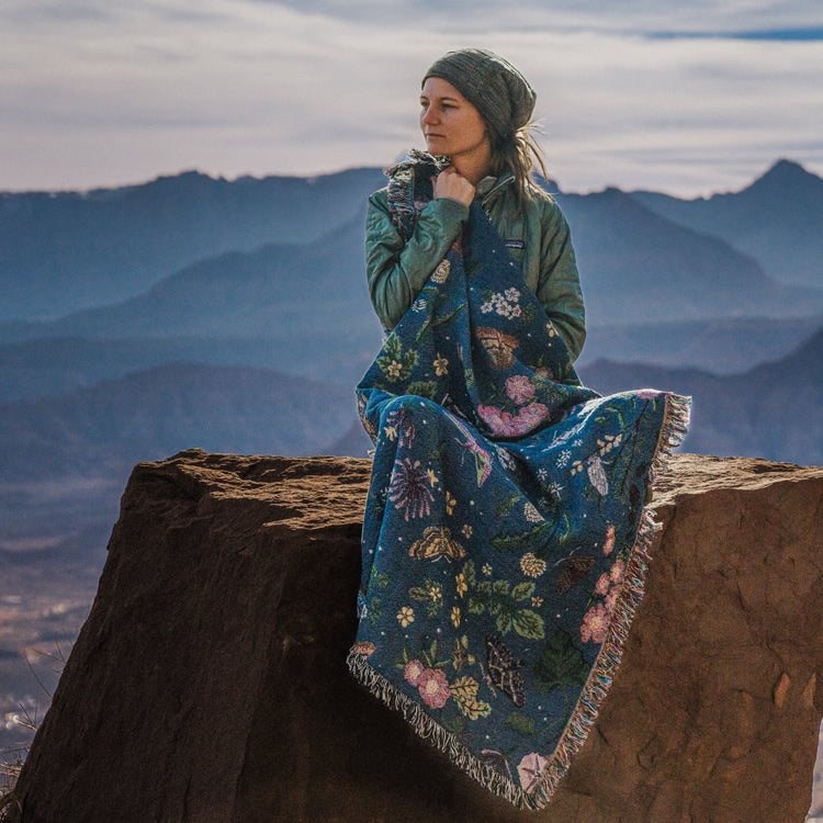 small dark blue woven wrap blanket with delicate moth and flower pattern around the shoulders of a woman in the mountains