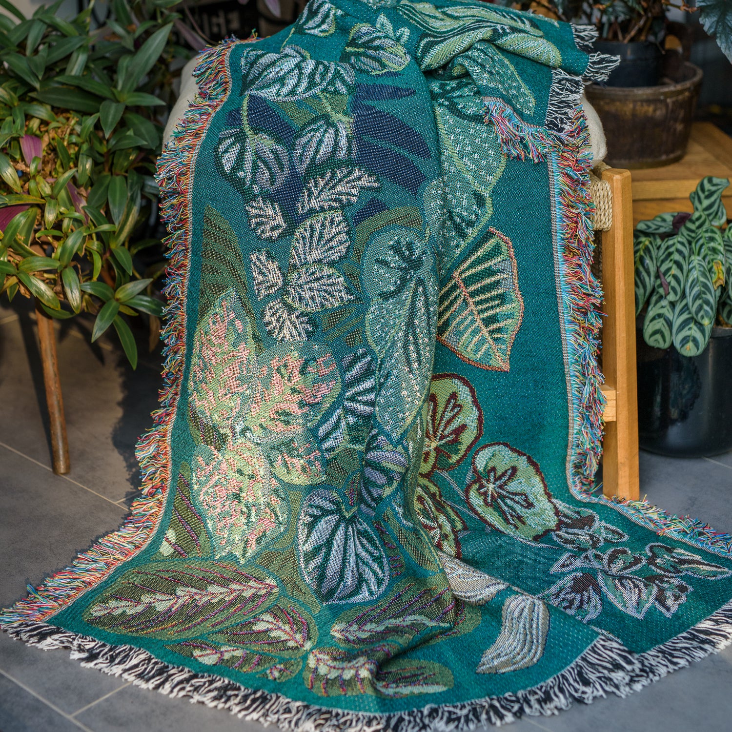 woven wrap blanket with fringe featuring lots of different houseplants draped over stool surrounded by houseplants