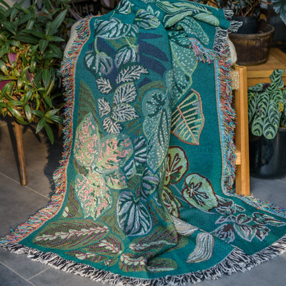 woven wrap blanket with fringe featuring lots of different houseplants draped over stool surrounded by houseplants