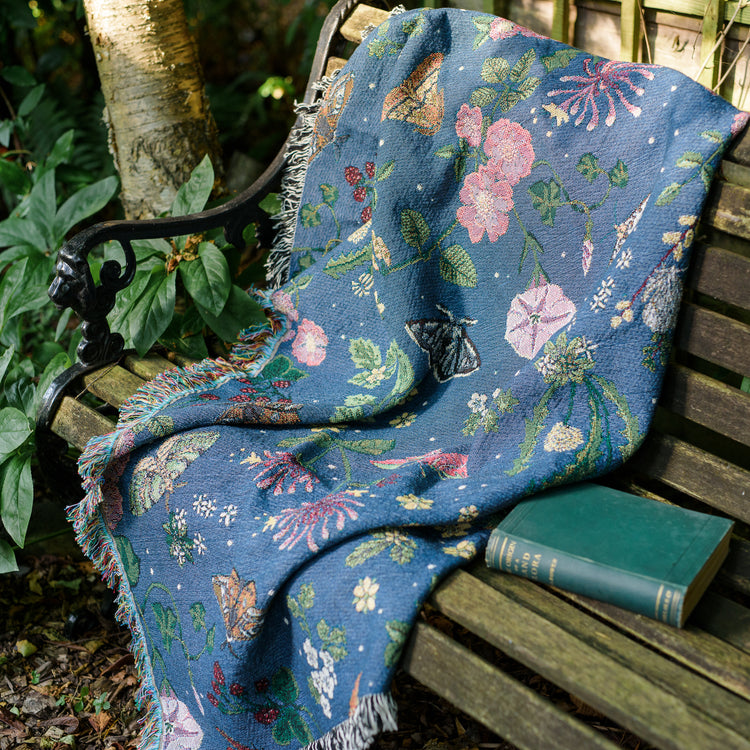 small dark blue woven wrap blanket with delicate moth and flower pattern on a bench in a garden with a book