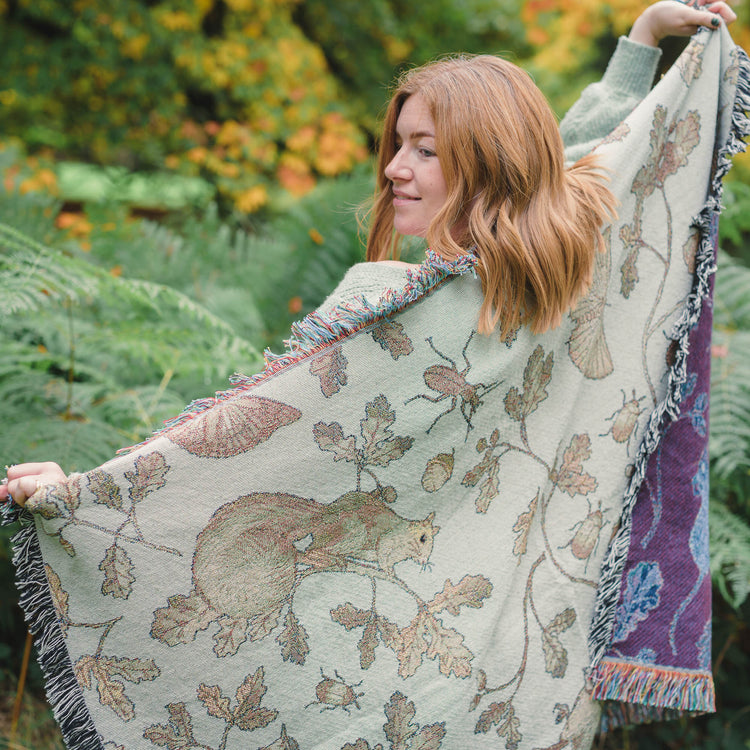 A woman wearing a Wild Oak Wrap Blanket in a forest, designed by Arcana.
